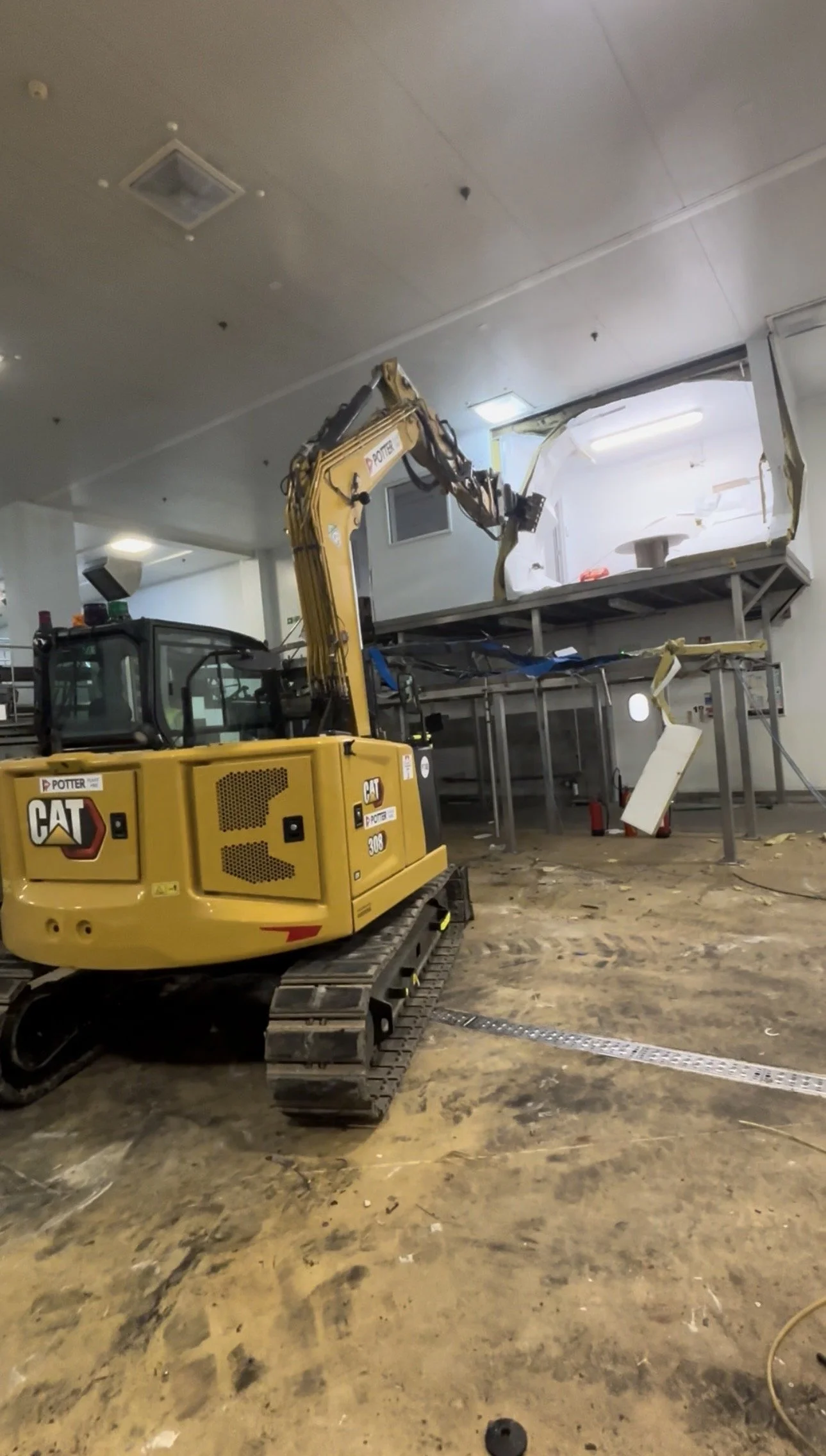 A yellow CAT excavator is demolishing a section of building, with debris and dust on the floor.