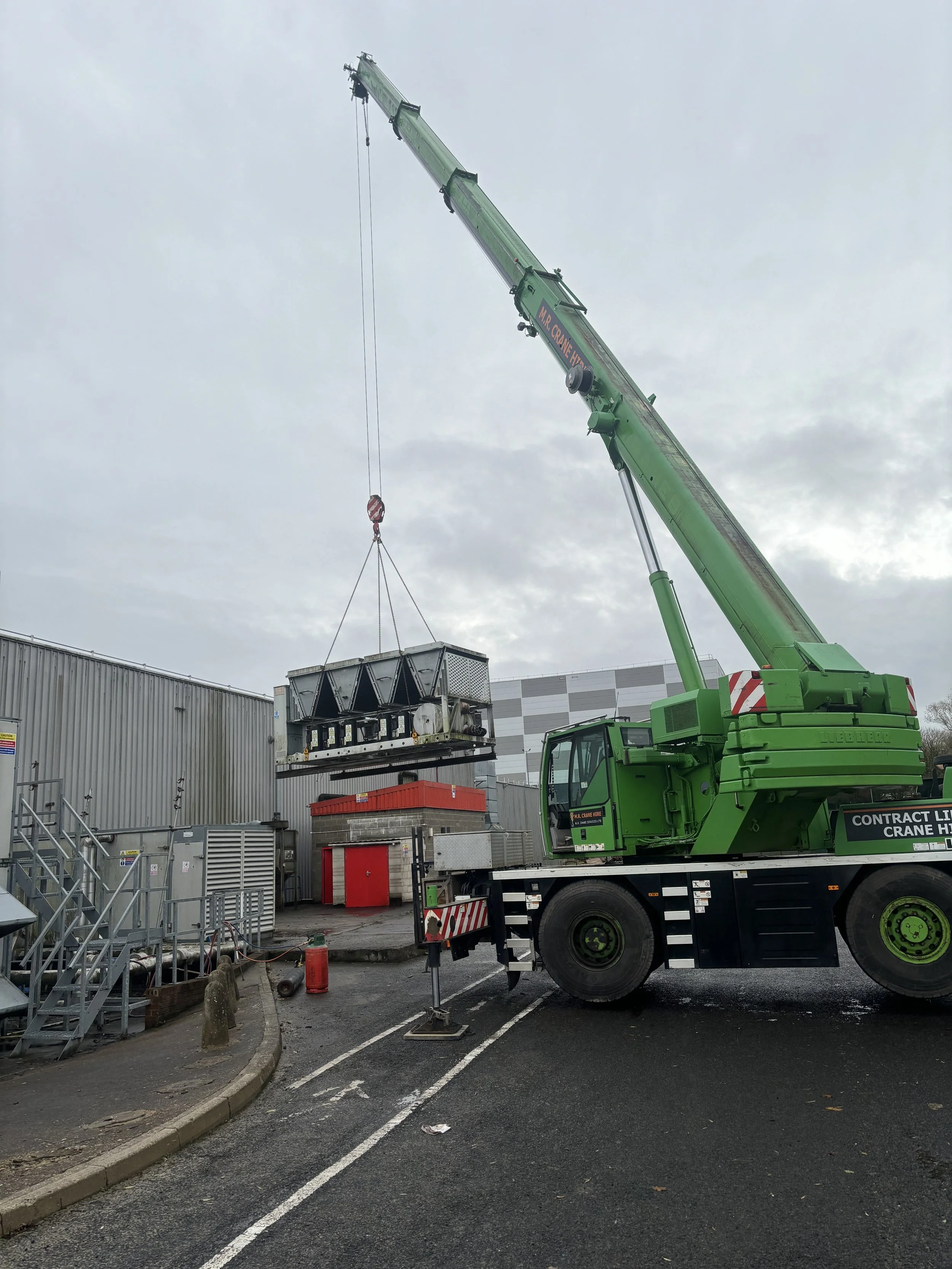 A large green crane lifting a water chiller at a food site with gray overcast sky.