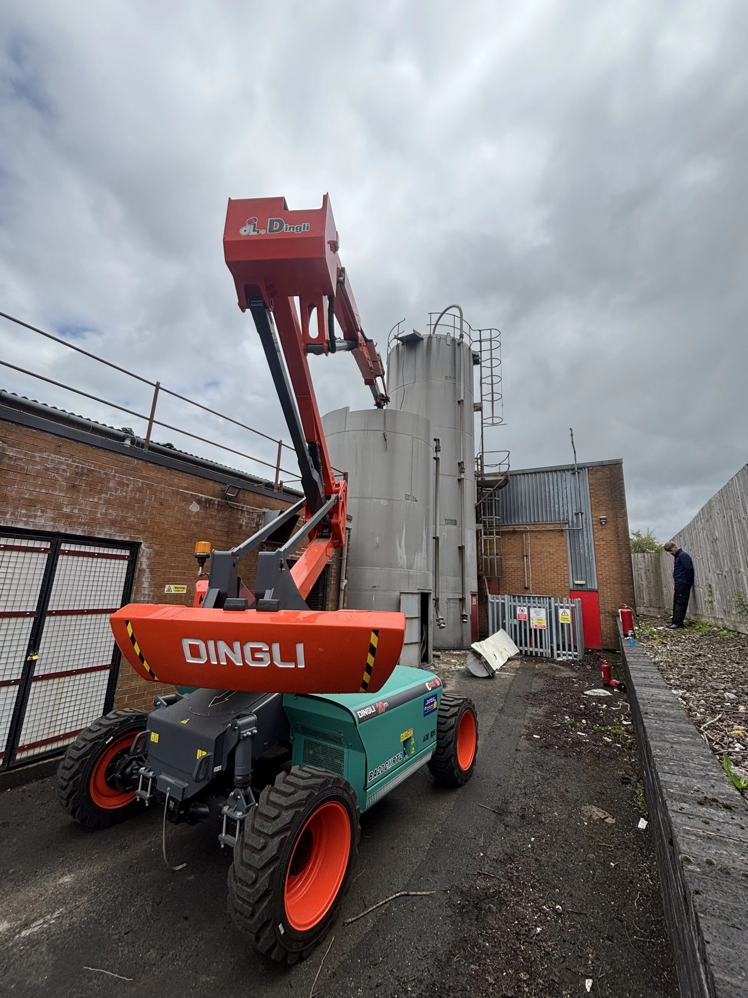 A red and green boom lift with the brand name 'DINGLI' on it, positioned near a brick building and industrial silos, with cloudy sky overhead and a person standing in the background.