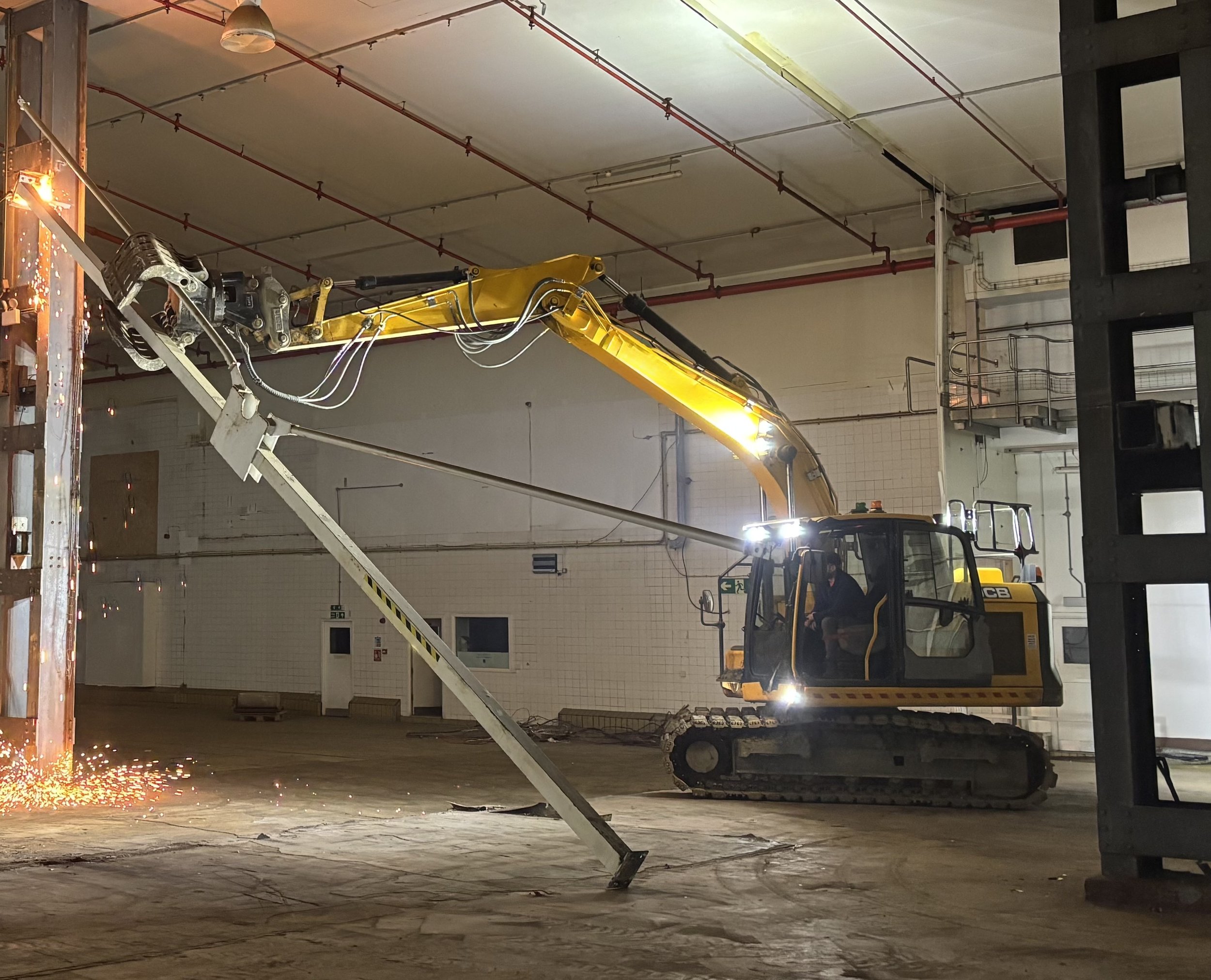 A construction site with a yellow excavator and a worker welding metal beams, sparks flying, inside a large industrial building with white tiled walls and exposed red pipes.