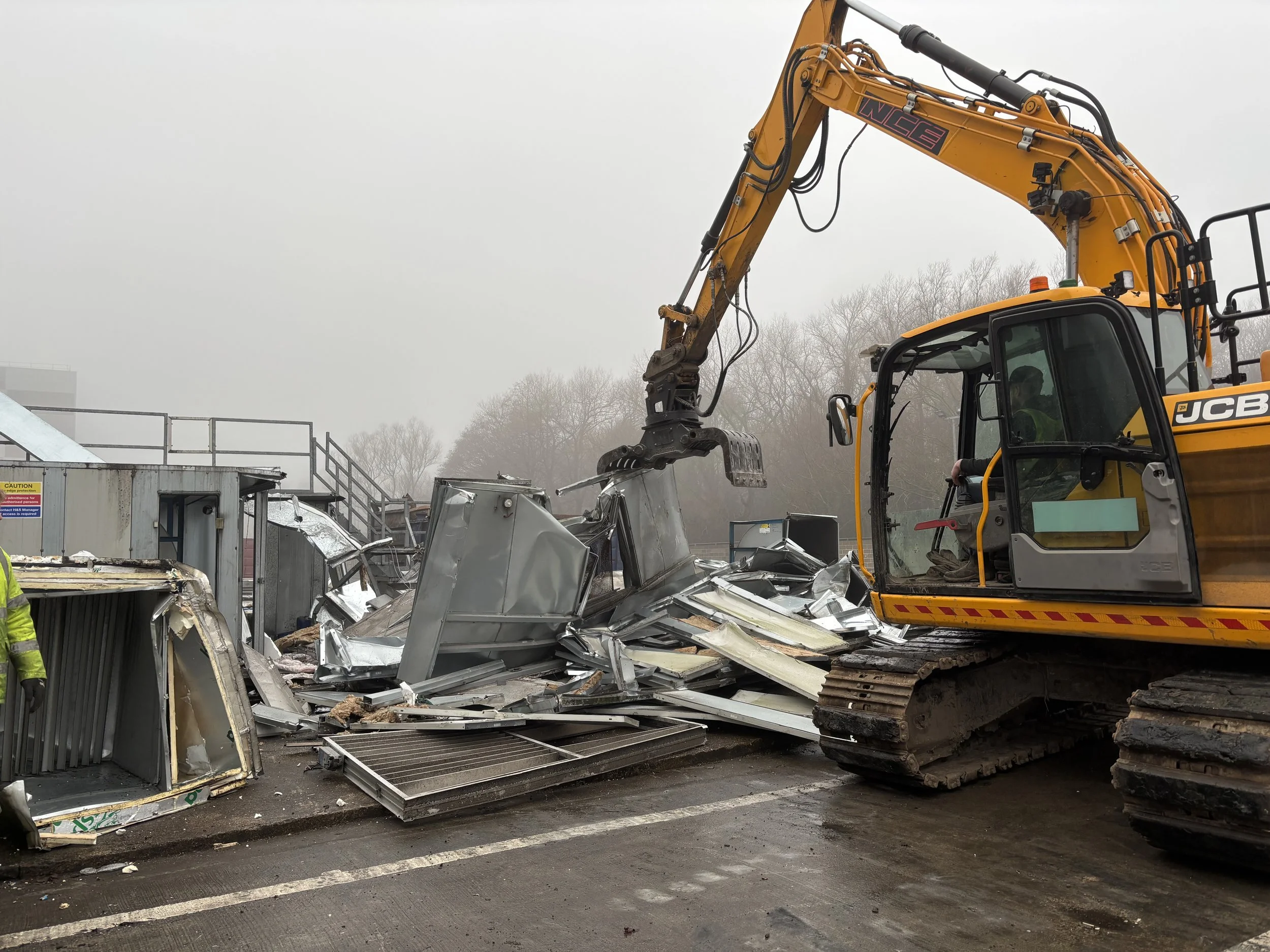 A large yellow excavator with the brand name JCB is demolishing a metal structure, with debris scattered around on the ground, in an outdoor area with a foggy sky and some trees in the background.