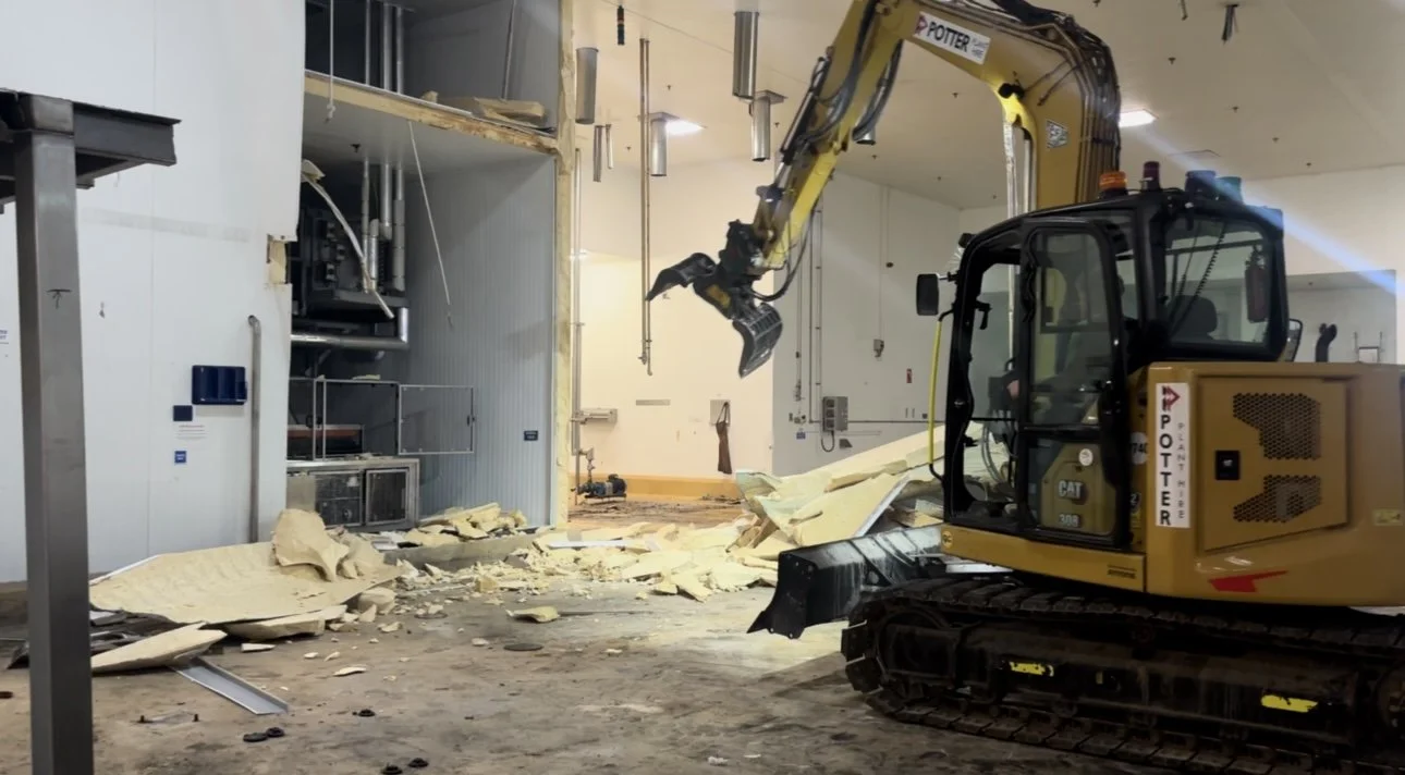 Interior stripout with a yellow Caterpillar excavator moving debris and insulation panels inside a food factory.