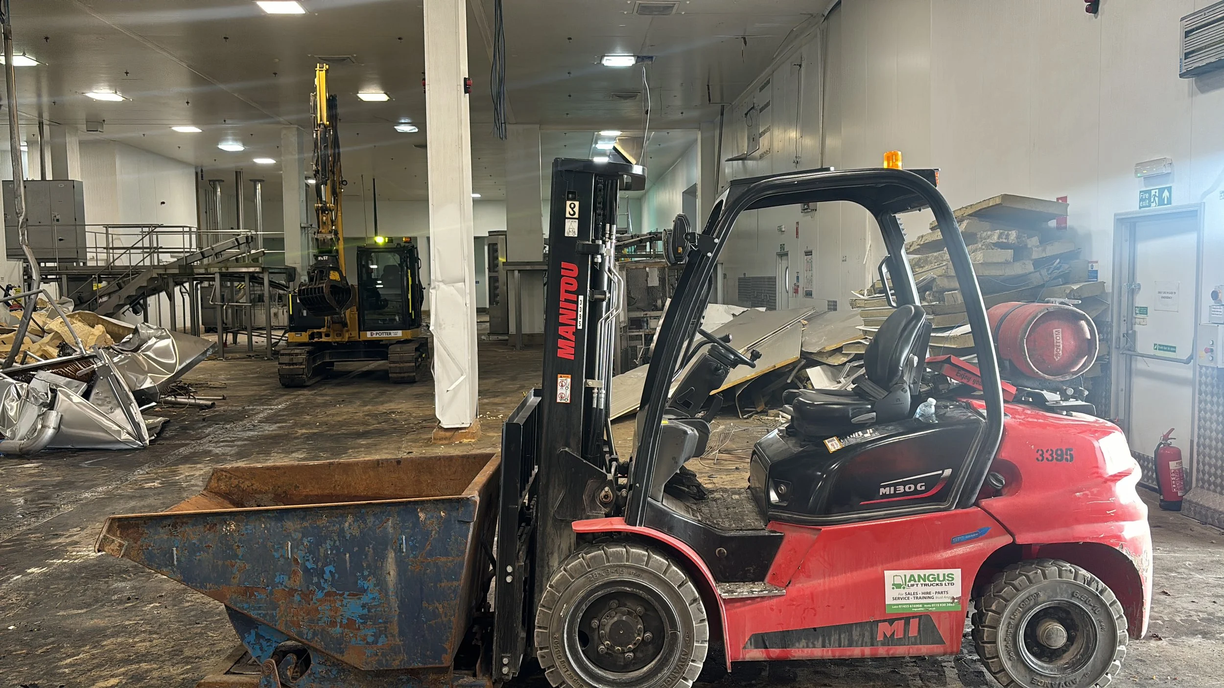 Indoor construction site with a red forklift in the foreground, and two small yellow construction vehicles in the background surrounded by debris and construction materials.