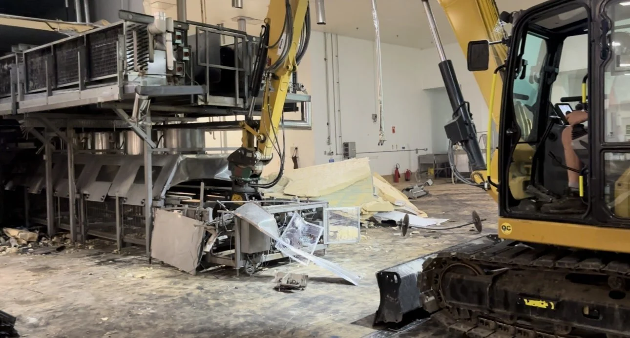 Construction equipment including a yellow excavator inside a building, with a partially demolished metal structure and debris on the floor.