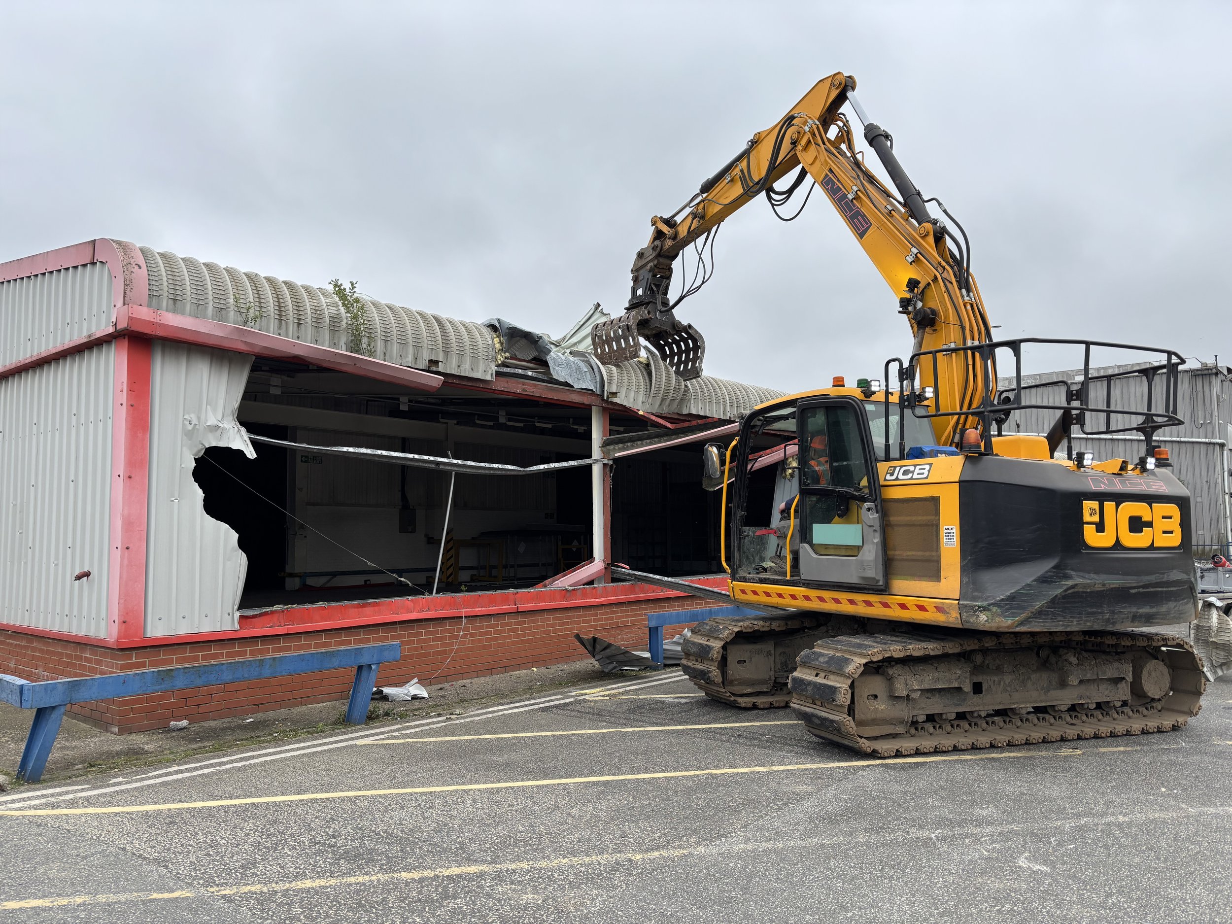 A construction site with a yellow excavator tearing down a building with a red and white corrugated metal exterior. The building's roof is partially collapsed, and debris hangs from the edges. The sky is overcast.