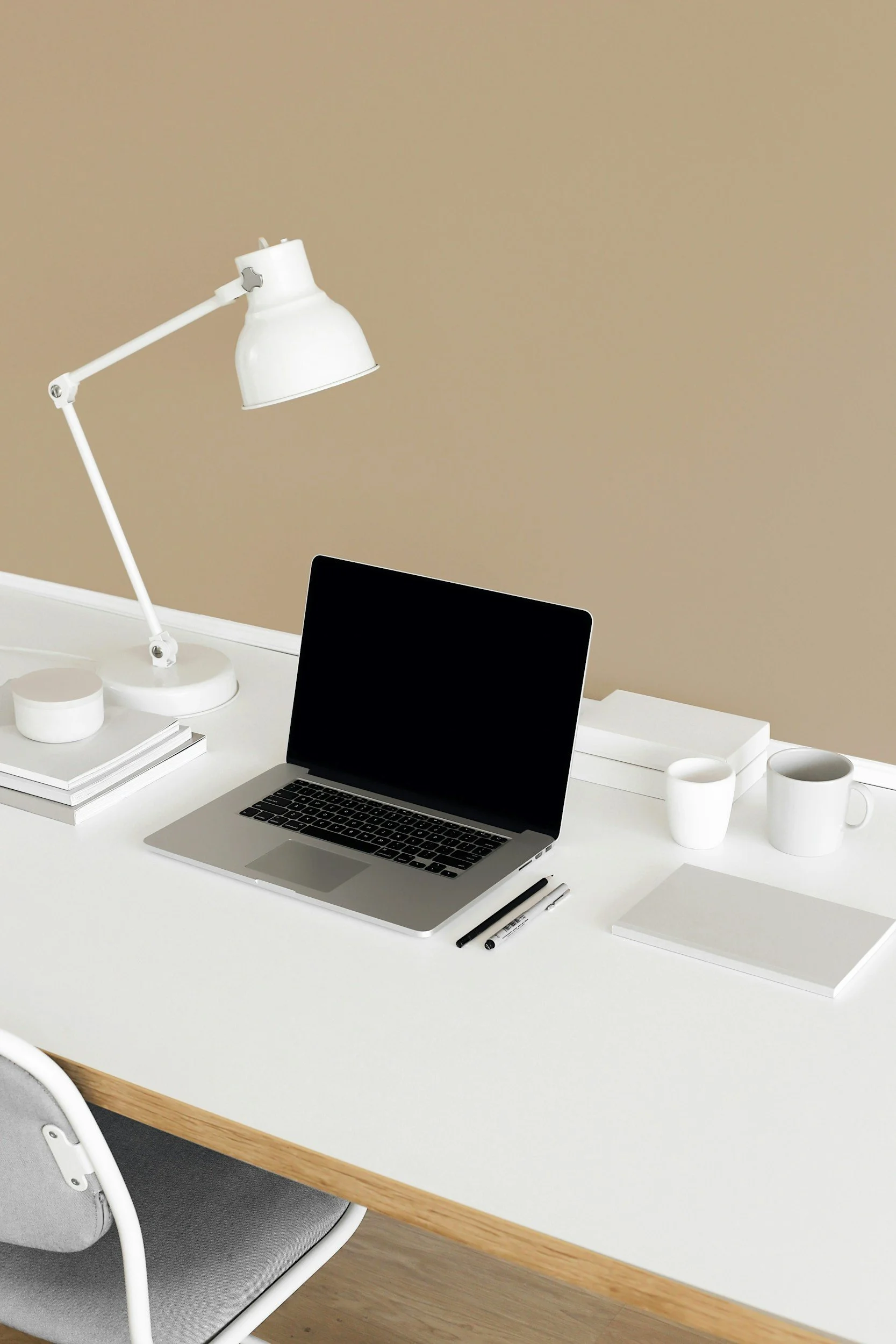 A minimalist white desk with a silver laptop, white coffee mugs, open notebooks, pens, and a white adjustable desk lamp against a beige wall.