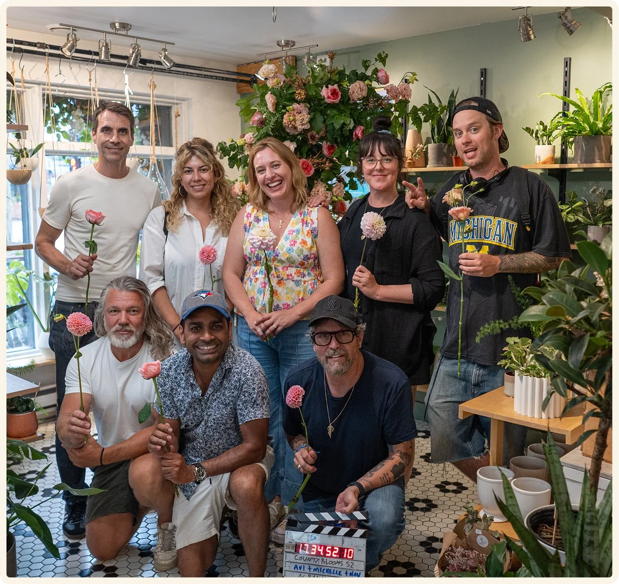 Group of nine people in a floral shop, holding flowers, with plants and flowers around them, and a film clapperboard on the floor.