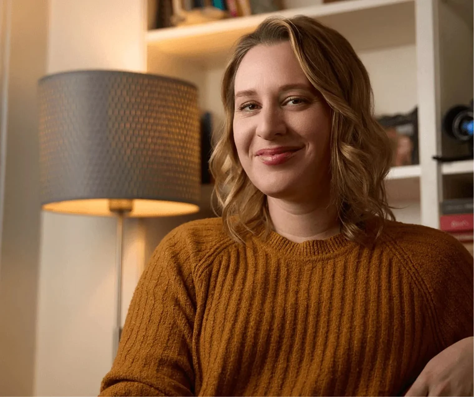 A woman with wavy blonde hair smiling while sitting in a room with a bookshelf and a table lamp.