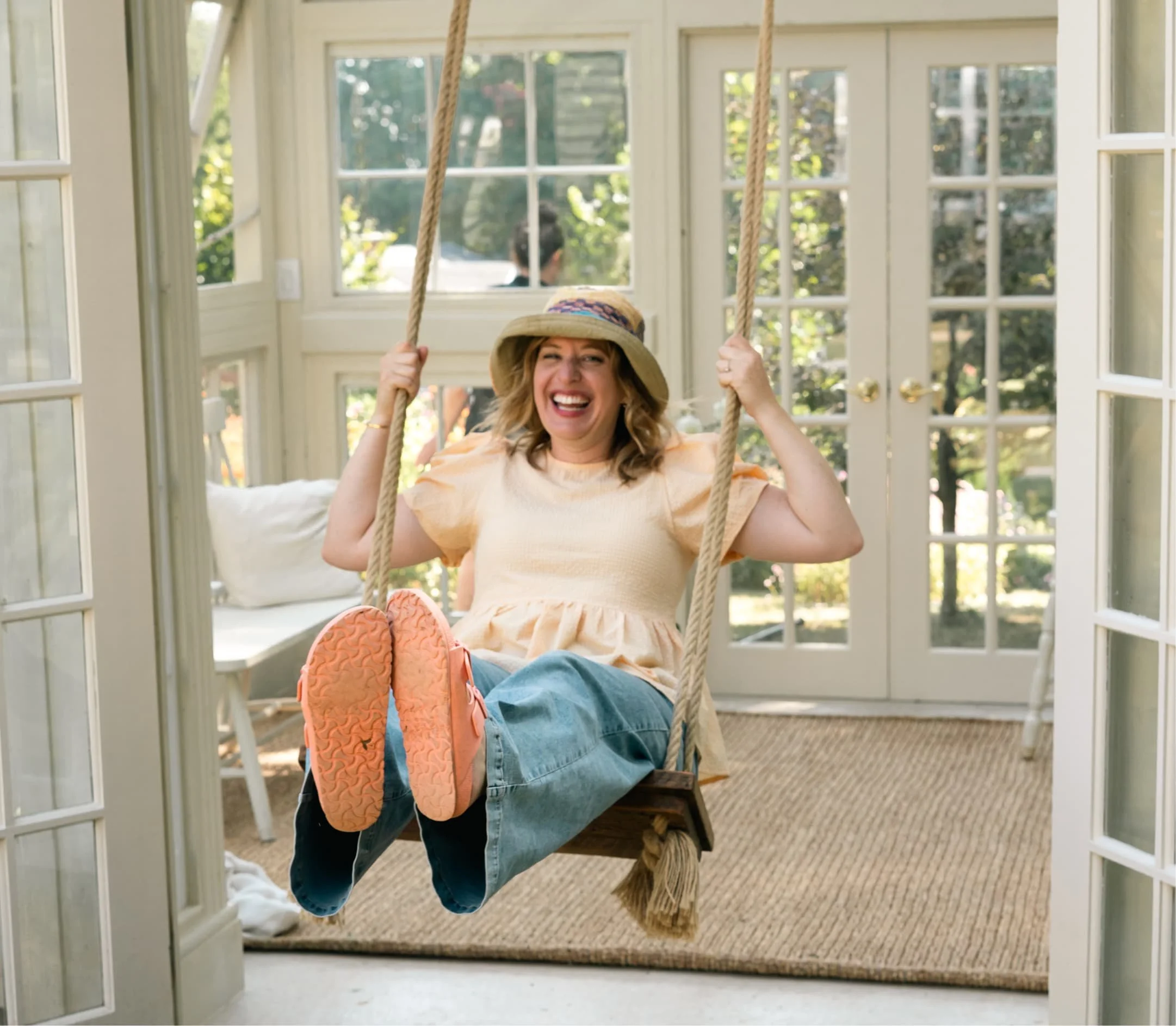 Woman smiling and sitting on a swing inside a sunroom with large windows and open glass doors, wearing a sunhat, yellow top, jeans, and pink sneakers.