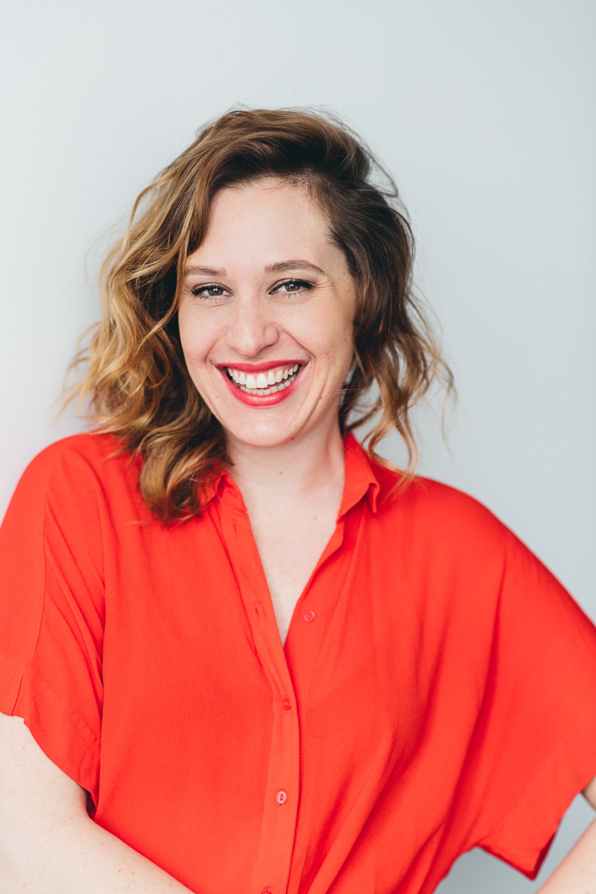 A woman with wavy, shoulder-length hair, wearing a bright red blouse, smiling with her teeth visible, against a plain light blue background.