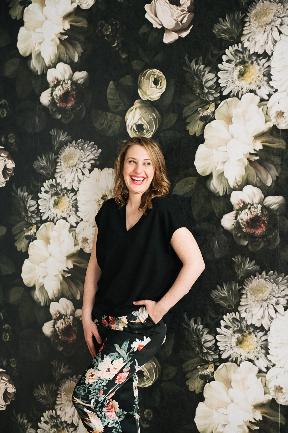 A woman with shoulder-length light brown hair, wearing a black top and floral pants, standing and smiling in front of a dark floral wallpaper with large white and cream flowers.