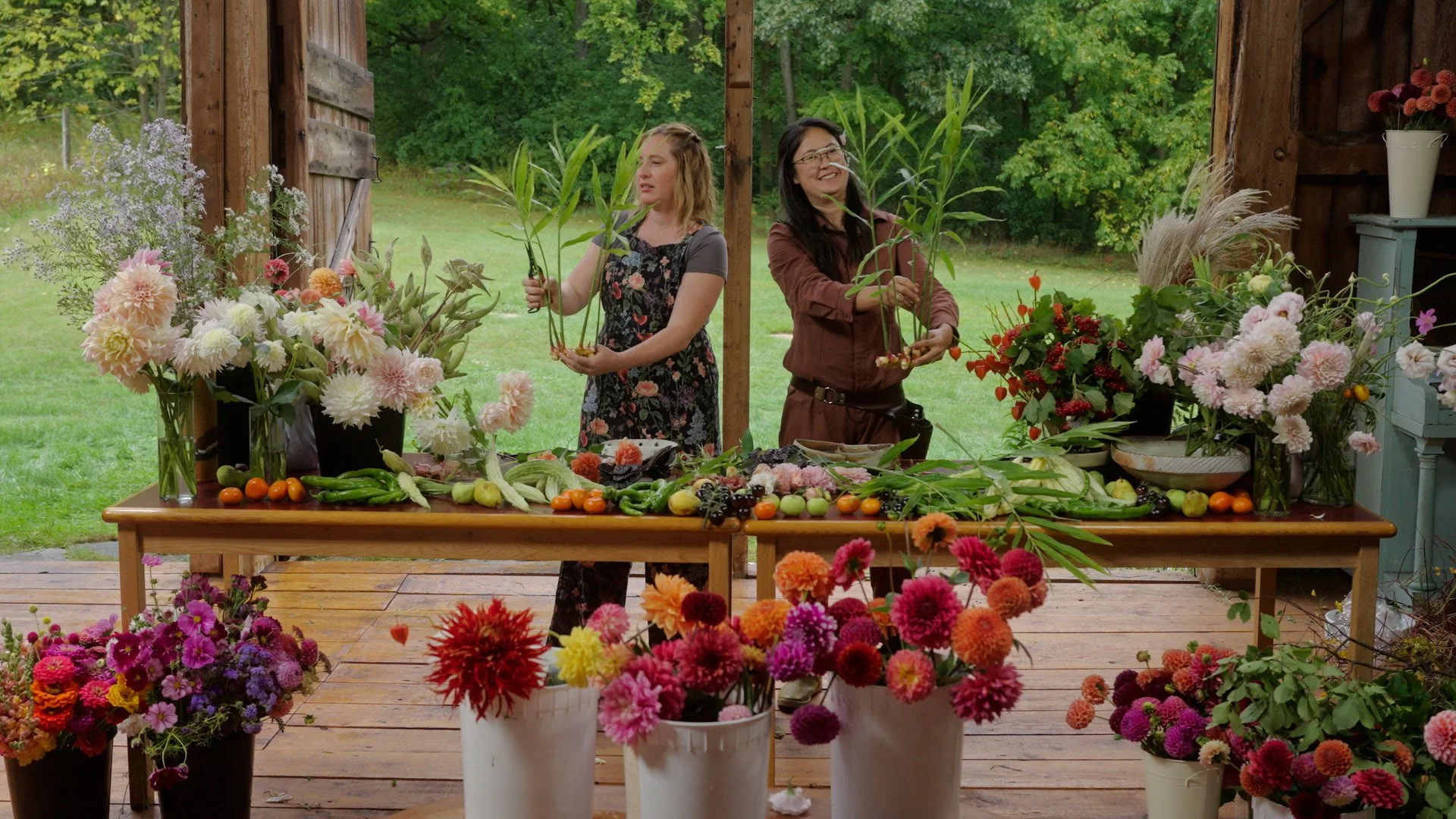Two women arranging flowers behind a wooden table with vegetables and flowers, in an outdoor setting with greenery.