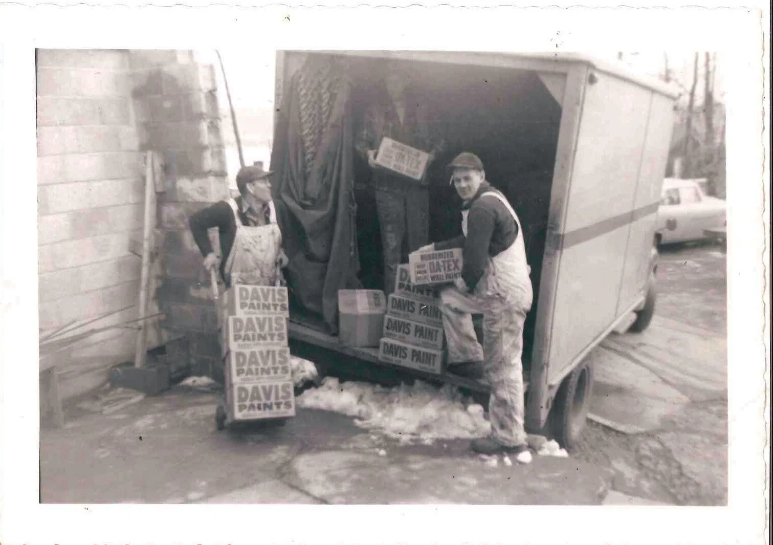 Two men in work uniforms unloading boxes labeled "Davis Paint" from a small delivery truck. One man is standing inside the truck, while the other is standing outside, holding a box. The truck is parked on a snowy street near a brick wall, and there i