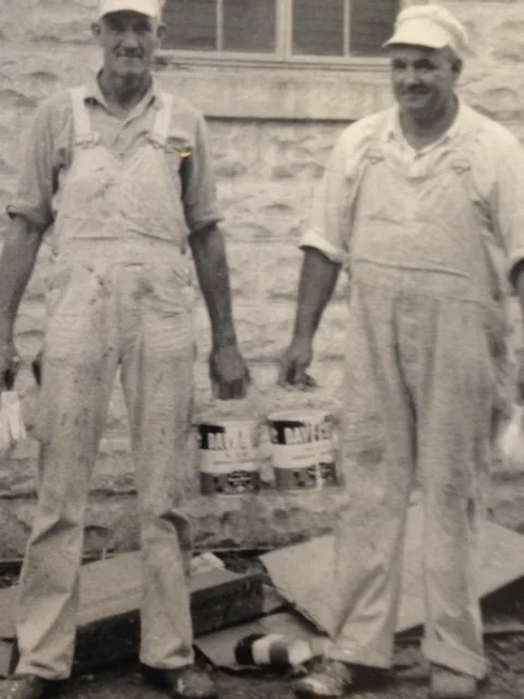 Two men in work overalls standing outside a building, holding cans of paint and a paintbrush.