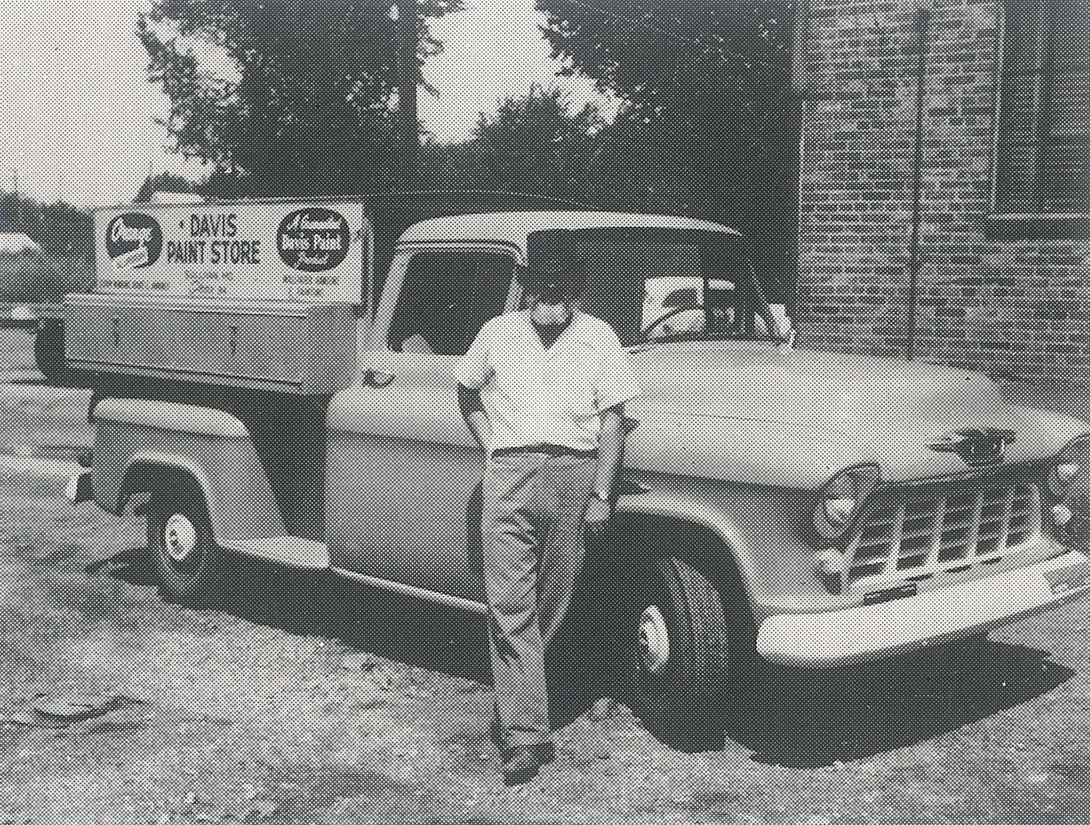 A person standing next to a vintage pickup truck with a sign for 'Lewis Paint Store' on the side, parked on a dirt surface with trees and buildings in the background.