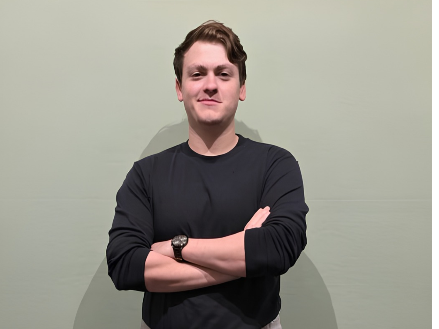 A young man with brown hair, wearing a black long-sleeve shirt and a watch, standing with arms crossed against a plain light green wall.