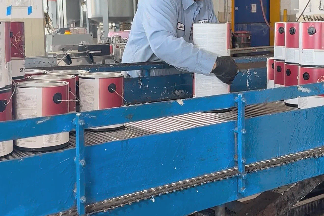 A worker in a factory filling white and pink cans with a product on a blue conveyor belt.