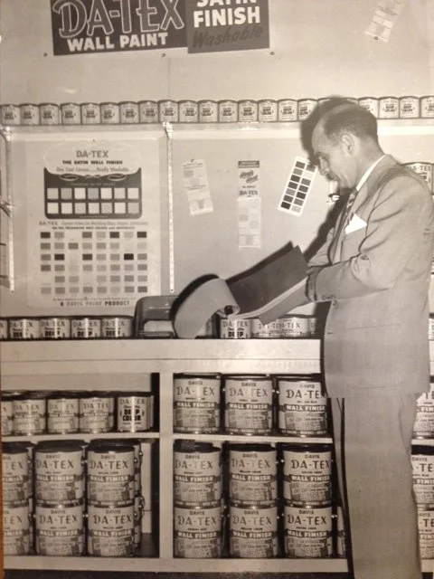 A man in a suit using a paint sprayer at a paint store, with paint cans and color swatches visible in the background.