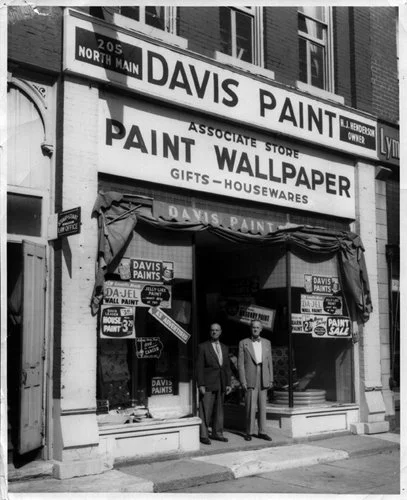 Black and white photo of the exterior of Davis Paint store with two men standing at the entrance, signage advertising paint, wallpaper, gifts, and housewares, located at 205 North Main street.