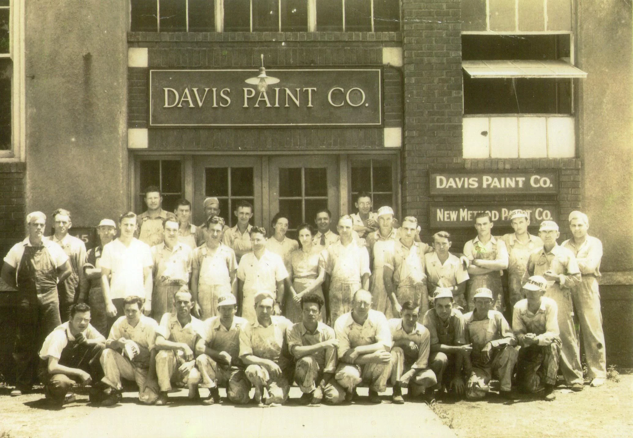 Black and white photograph of a large group of men standing outside a building with signs reading 'Davis Paint Co.' and 'New Met.' The men are dressed in work clothes and uniforms, some wearing caps. The building has large windows and a brick facade, with an industrial or commercial appearance.