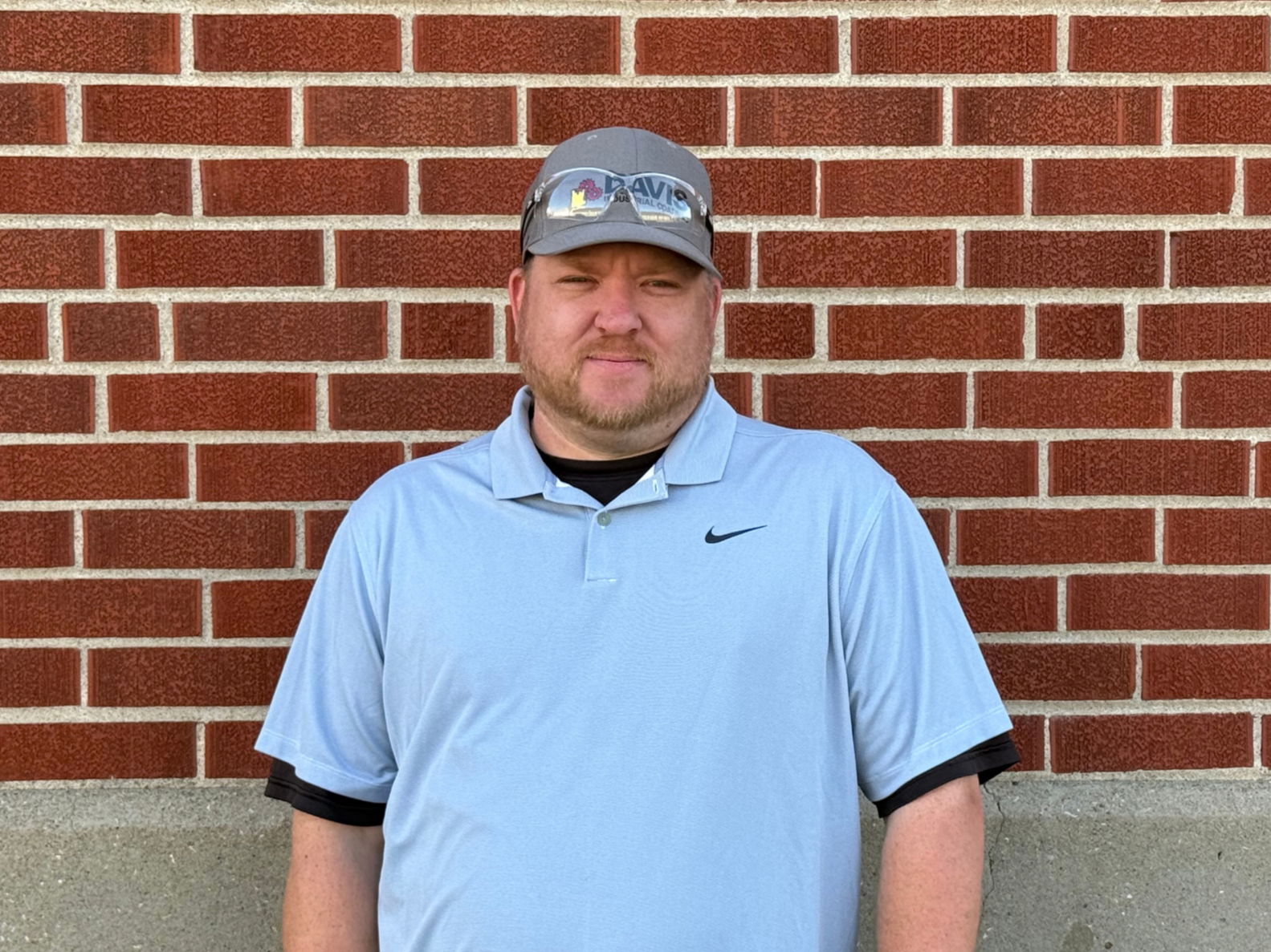A man standing outdoors in front of a red brick wall, wearing a light blue polo shirt, a gray baseball cap with a logo, and glasses. He has a beard and is looking at the camera.
