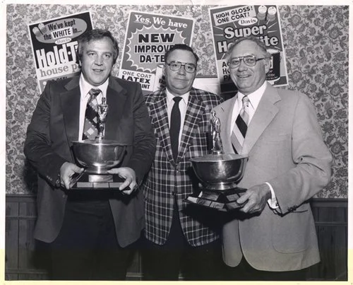 Three men in suits and ties standing indoors, holding trophies, smiling, with political campaign posters on the wall behind them.