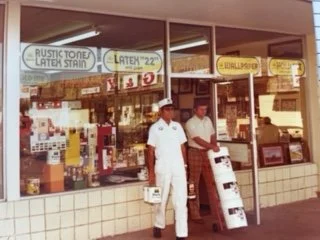 Two men standing outside a storefront with signs advertising rustic stone after stain, latex paint, and drywall supplies. One man is dressed in white, the other in a light shirt. The storefront has large windows and various products inside.