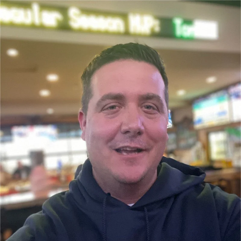 A man smiling in a casual dining restaurant or food court, with blurred background of digital menu screens and other people.