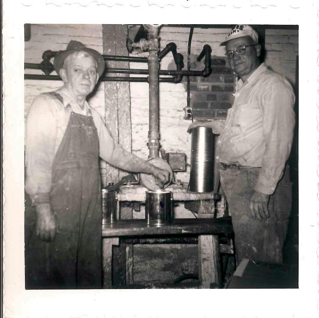 Two people working with metal cylinders and tools in an industrial setting, one woman wearing overalls and a hat, and one man in a hat and glasses.