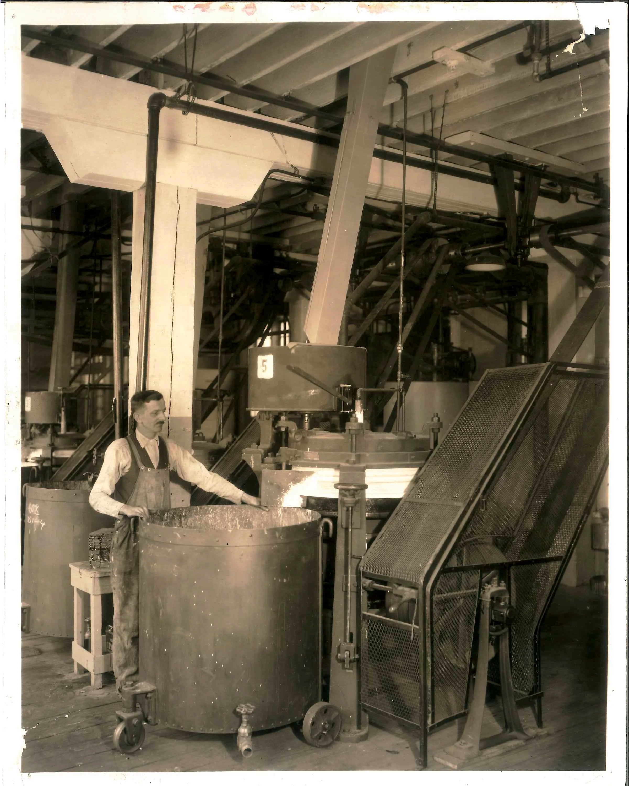 Industrial scene with machinery, pipes, and a man standing next to a large metal container or drum on wheels. The background shows a ceiling with beams and various industrial components.