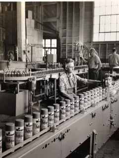 A person working at a manufacturing or packaging line with cans and bottles, in an industrial setting with high ceilings and large windows.
