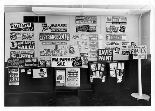 Wall display with multiple signs advertising wallpaper and paint sales, including clearance and discount offers, on a black wall with some hanging samples and smaller informational posters.