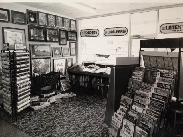 Interior of an art supply store with framed artwork on the wall, shelves of art books, and display racks of craft supplies and papers.