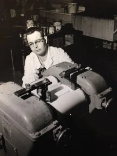 A woman wearing glasses working with a machine in a laboratory or workshop setting, with shelves and containers in the background.