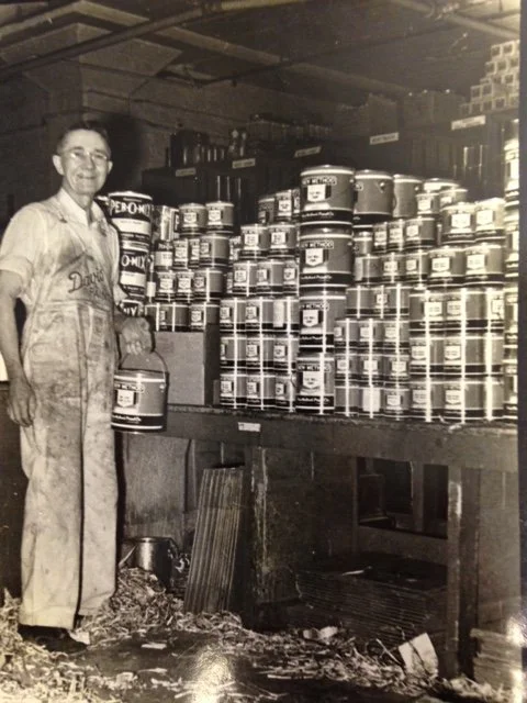 A woman in work overalls standing next to shelves filled with cans of paint in a workshop or storage room, black and white photo.
