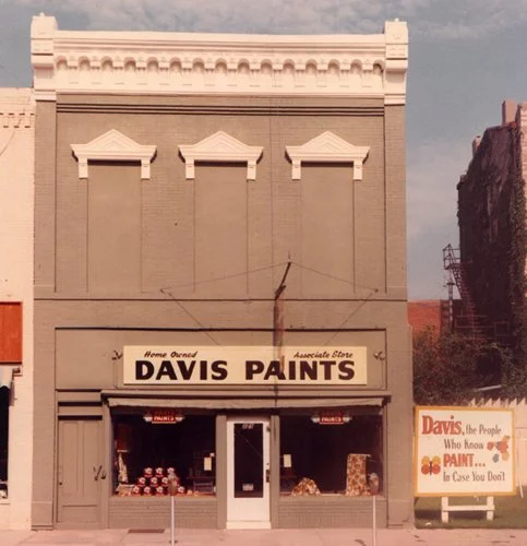 A two-story building with a paint store called Davis Paints on the ground floor. The building is tan with decorative white trim at the top, and there are three triangular decorative elements above the store sign. Outside the store, various paint cans
