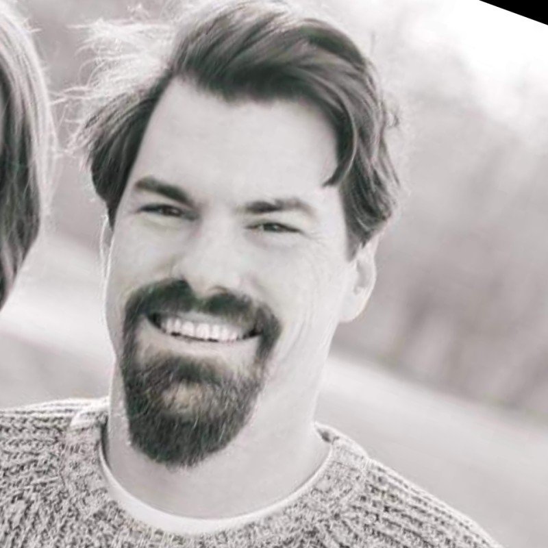 A smiling man with dark hair, a beard, and a mustache, wearing a knit sweater, outdoors in a natural setting.