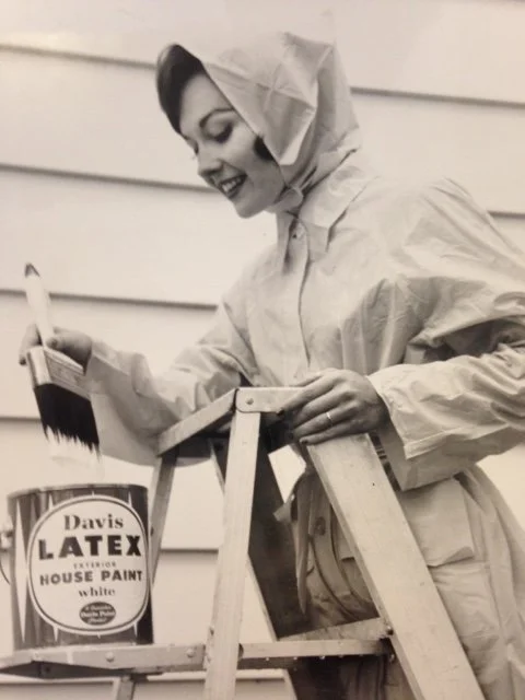 A woman in a hooded raincoat smiling while painting a wooden object outdoors. She is holding a paintbrush and a small tool, with a can of latex house paint nearby.