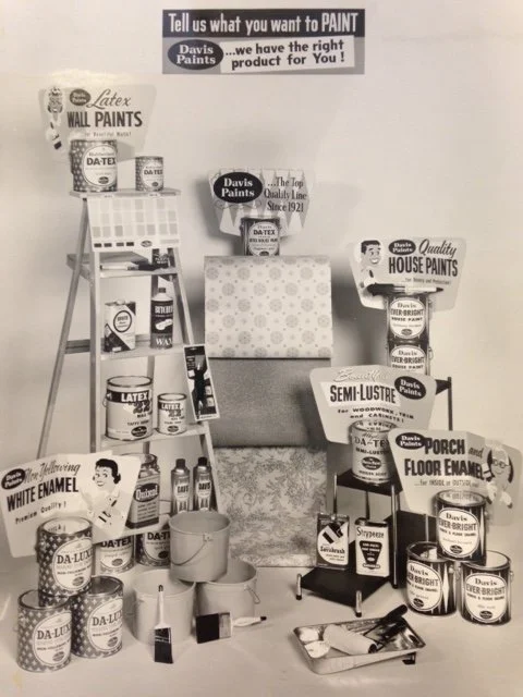 Display of various home paint products, including wall paints, house paints, semi-lustre, porch floor enamel, white enamel, and latex paints, along with paint brushes and rollers, arranged on shelves, a chair, and the floor in a promotional setting w