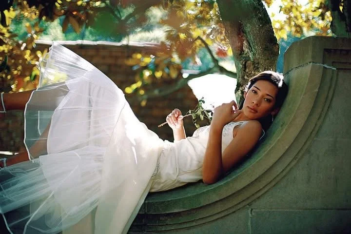 A woman in a white dress lying on a curved green stone bench outdoors, surrounded by trees and sunlight filtering through the leaves.