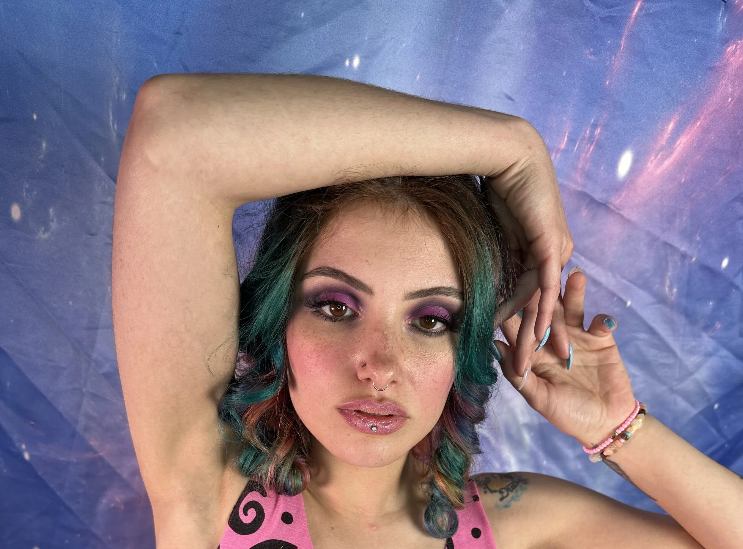 A woman with multi-colored hair, pink eyeshadow, and shiny pink lipstick, lying down against a purple background with galaxy patterns, with her left arm bent over her head, smiling.