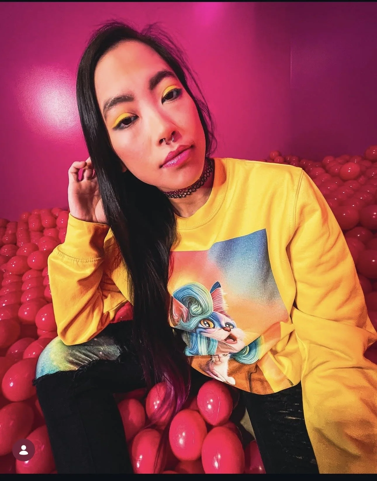 A young woman with long black hair and colorful makeup, wearing a yellow sweatshirt with a graphic of a fantasy creature, sitting against a pink background with red spherical objects.