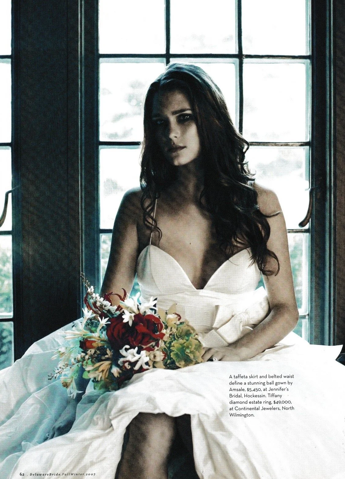 A woman with long dark hair and dark eye makeup sitting in front of a window, wearing a white gown with a taffeta skirt and belted waist, holding a bouquet of flowers.