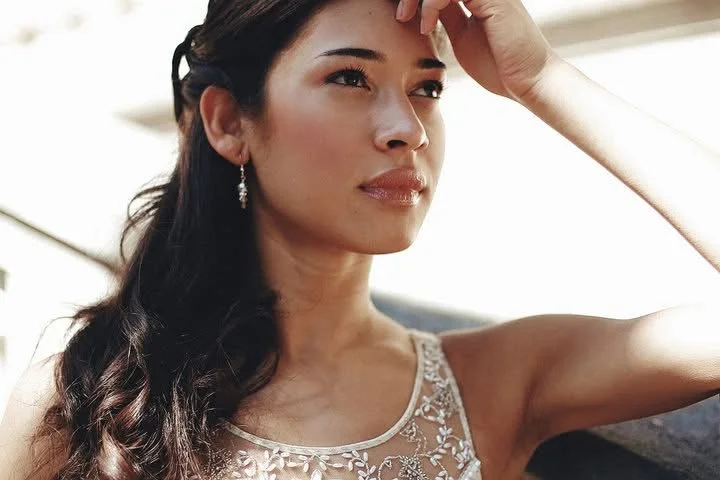 A woman with long dark hair styled in loose waves, wearing a sleeveless, lace-detailed dress, and earrings. She is touching her forehead with her right hand, looking thoughtful, with a light background.