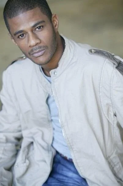 Portrait of a man with short hair and a serious expression, wearing a light-colored jacket over a blue shirt.