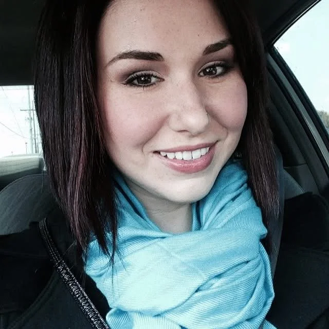 A young woman with dark hair, smiling, wearing makeup and a blue scarf inside a vehicle.