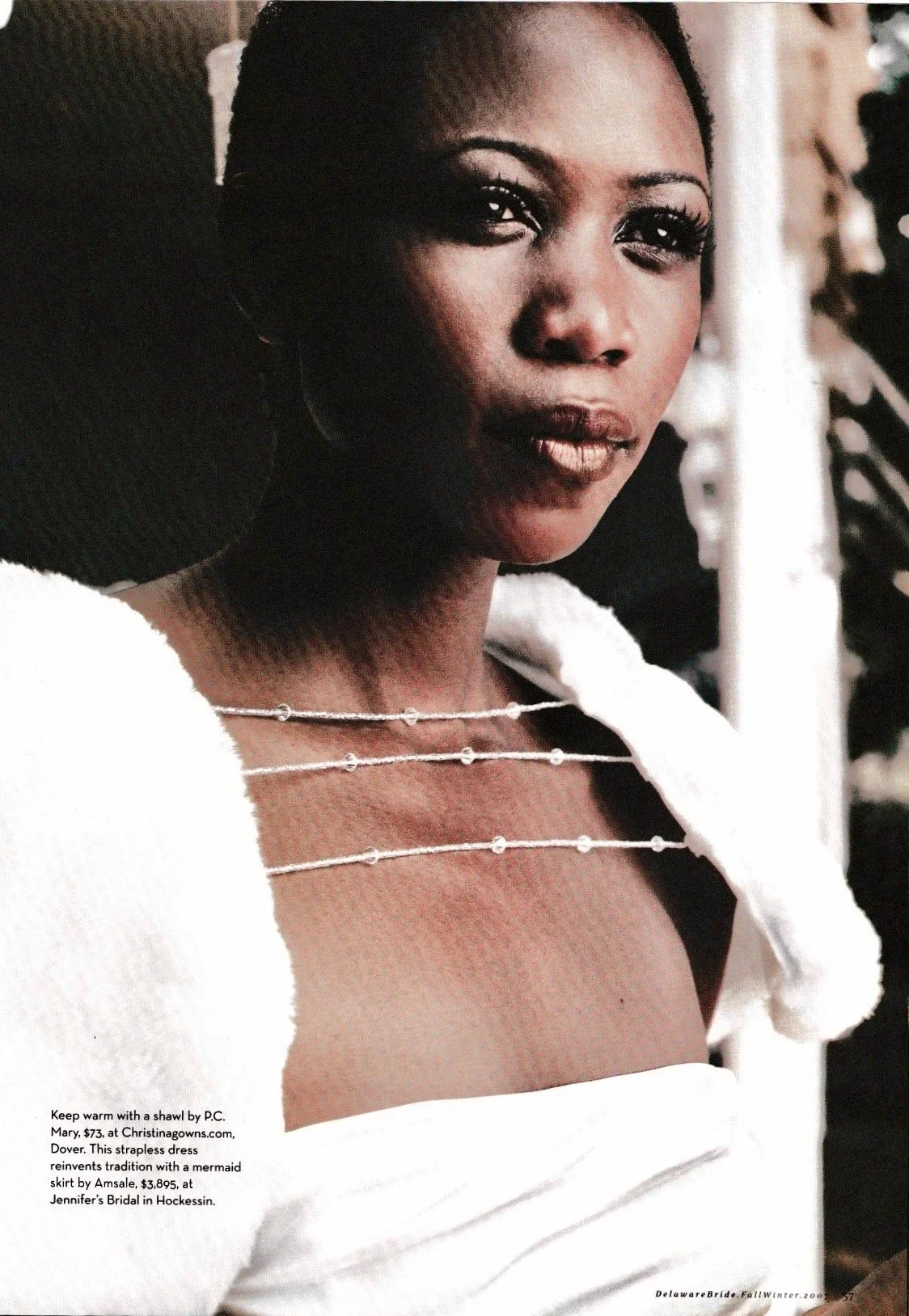 A woman with short hair, dark skin, and wearing makeup including eyeliner and lipstick. She is dressed in off-white and wearing layered delicate necklaces. The background is blurred and she is looking directly at the camera.