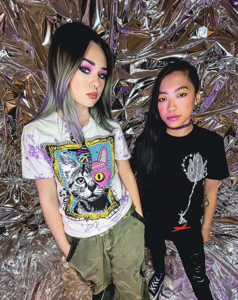 Two young women standing in front of a shiny, crinkled metallic backdrop. The woman on the left has gray and purple ombre hair and wears a white tie-dye t-shirt with a graphic of a cat's face and colorful design, along with khaki cargo pants. The wom