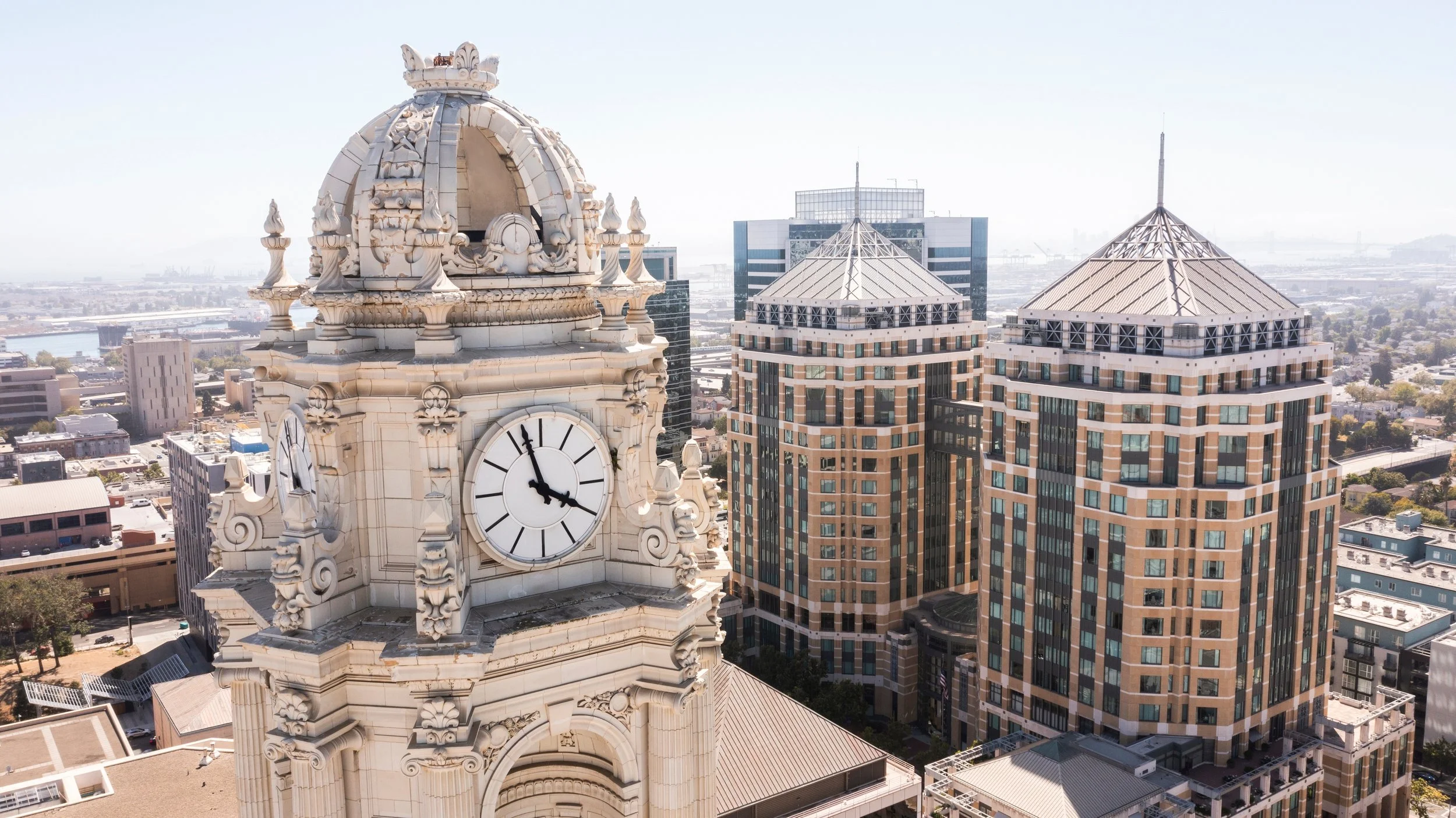 A cityscape featuring a historic clock tower with a white ornate dome and two newer high-rise buildings with pointed tops in an urban area, with a river and more city buildings in the background.