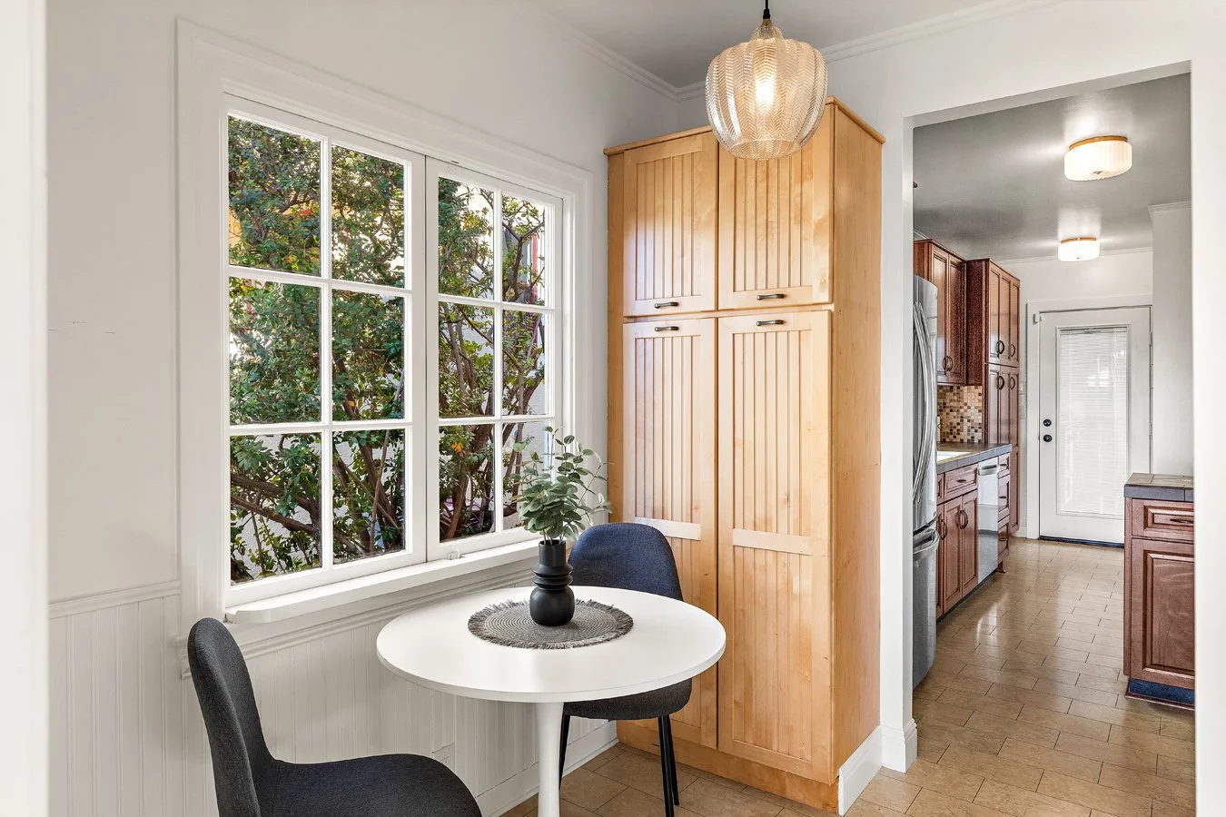 Kitchen dining area with a small round white table, two black chairs, a black vase with green plants, and a large window showing greenery outside. Wooden cabinets and a door leading outside are visible in the background.